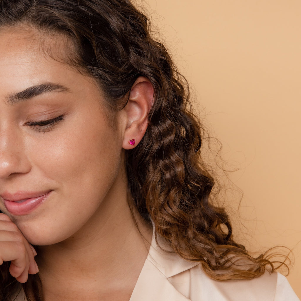 A woman with curly brown hair wears Ruby Heart Studs, delicately styled as she smiles softly against a beige background.