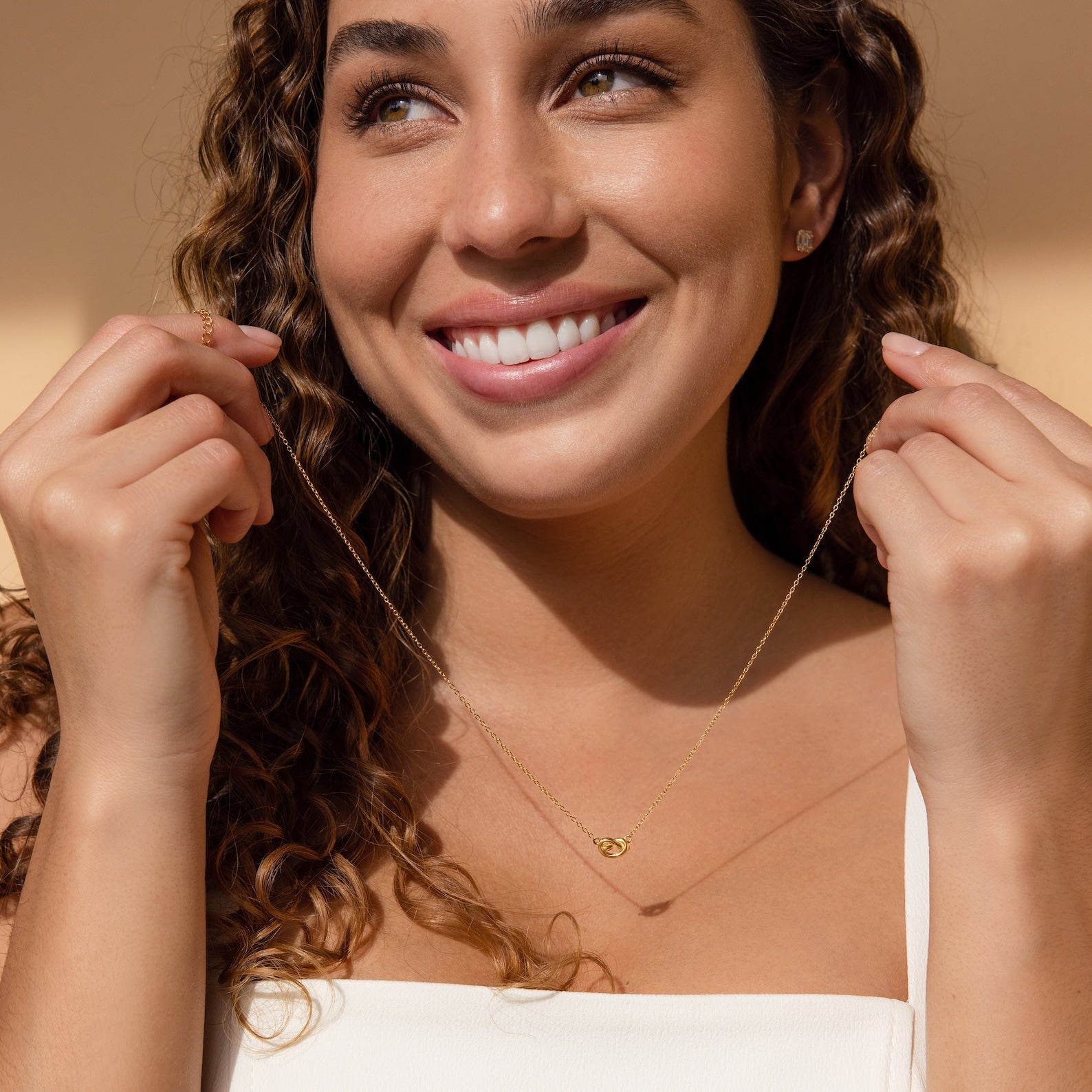 A smiling woman with curly hair, dressed in a white top and bathed in natural light, showcases the Love Knot Necklace—an ideal wedding gift from Unity Jewelry.