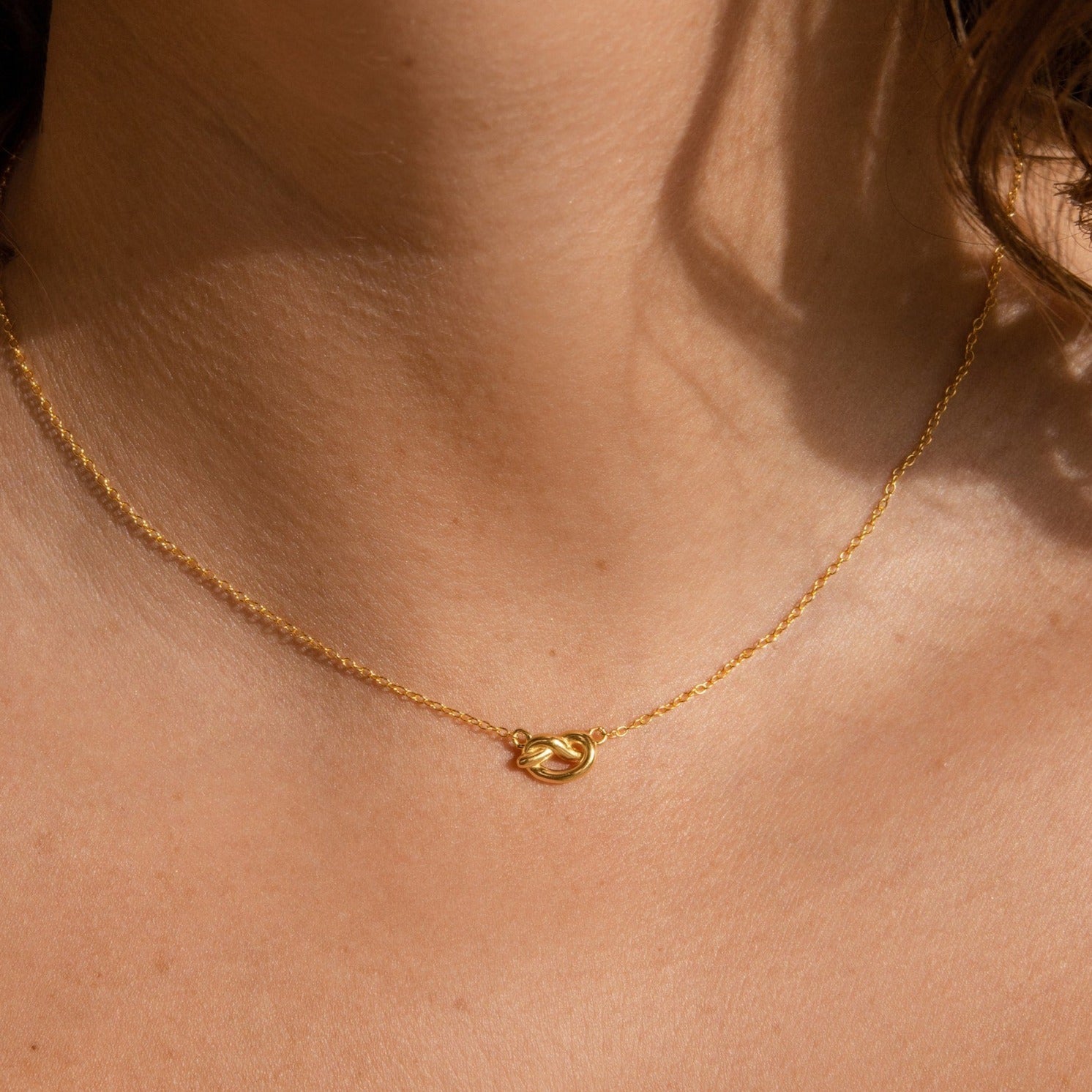 The Love Knot Necklace, a delicate gold piece featuring a small knot pendant, elegantly rests on the neck—an ideal wedding gift symbolizing unity and everlasting togetherness.