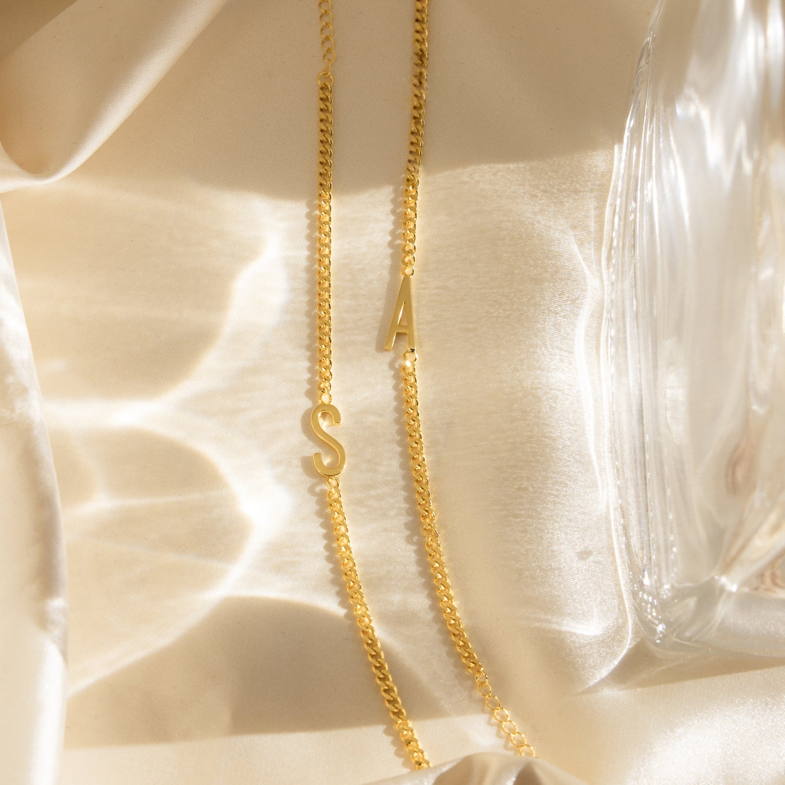 Large Letter Bracelets with S and A rest on silky cream fabric beside a glass item, ideal for pairing with your favorite everyday jewelry like a classic curb chain bracelet.
