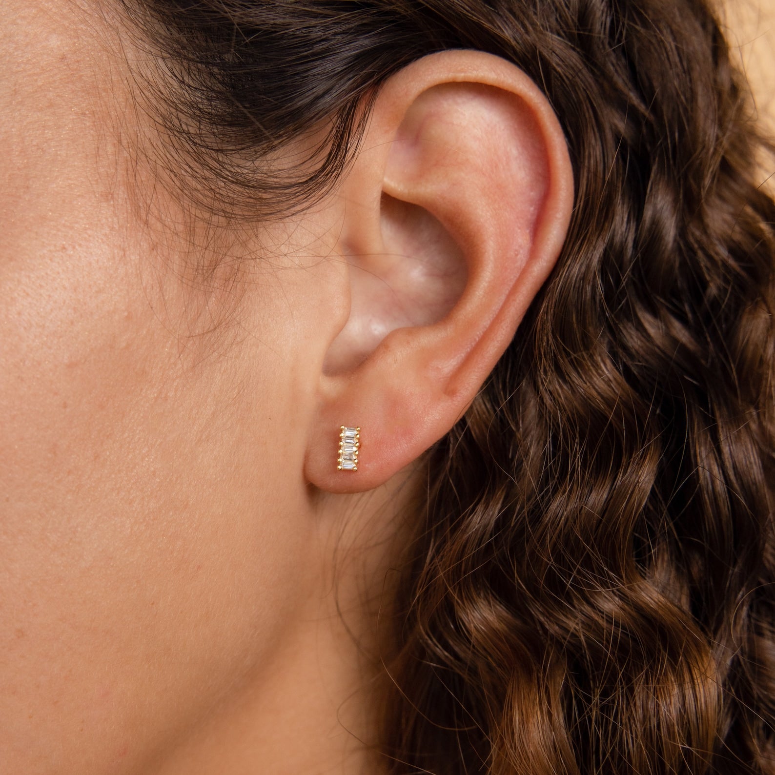 Close-up of a woman's ear with wavy brown hair, wearing Serena Baguette Studs—minimalist stud earrings featuring small gold and diamond details.