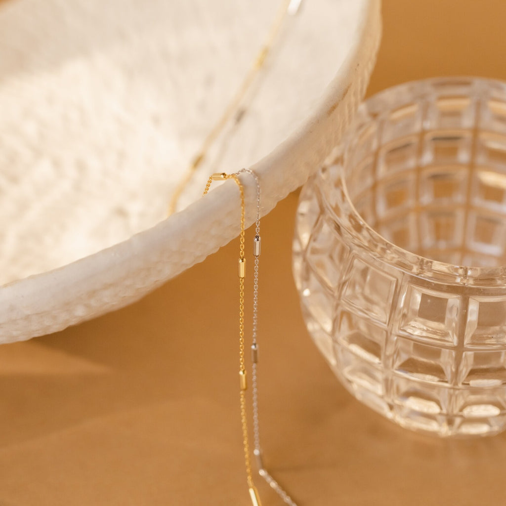 A Bar Station Necklace with delicate chains drapes over a textured white bowl, accented by dainty charms, beside a clear glass cup on a tan surface.