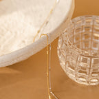 A Bar Station Necklace with delicate chains drapes over a textured white bowl, accented by dainty charms, beside a clear glass cup on a tan surface.