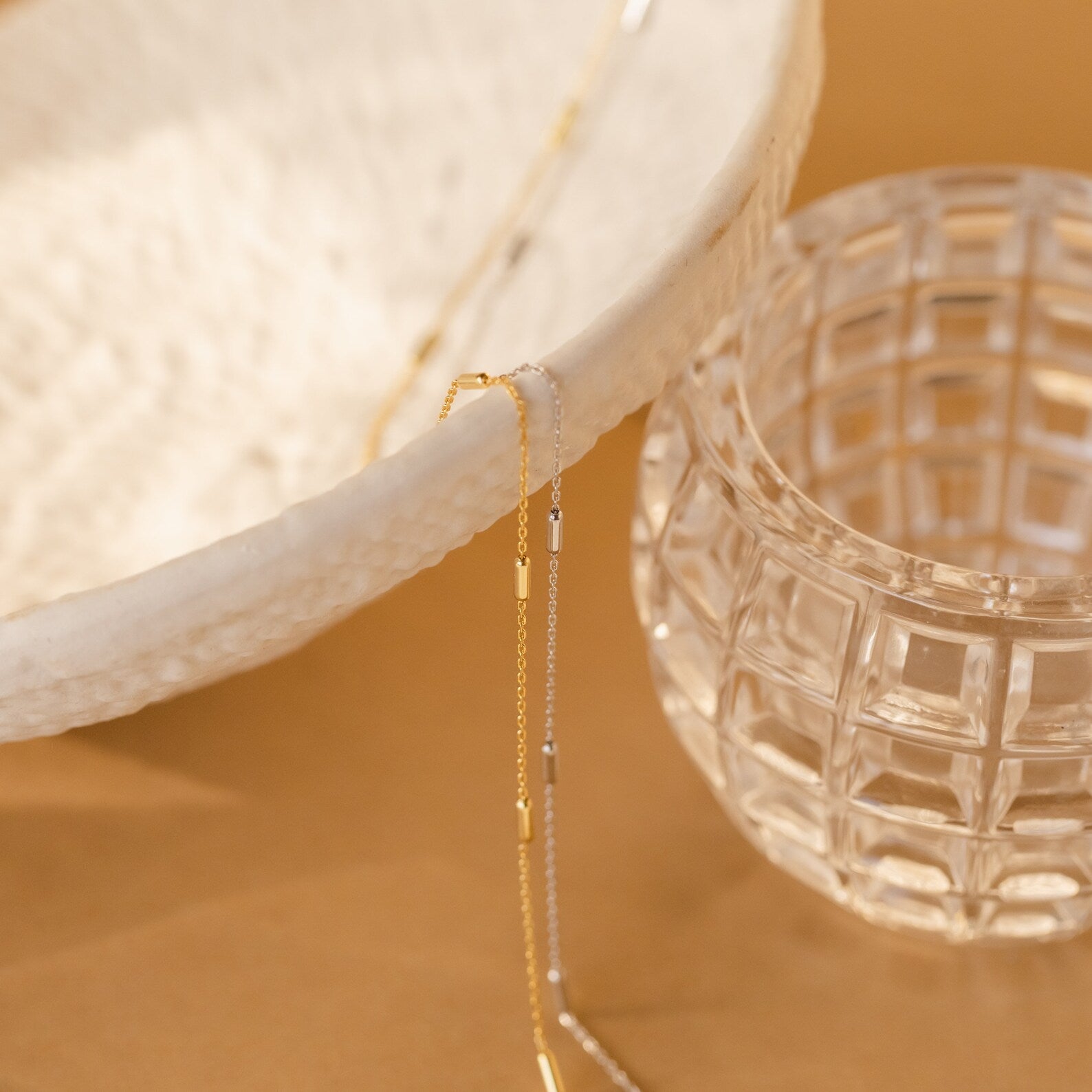 A Bar Station Necklace with delicate chains drapes over a textured white bowl, accented by dainty charms, beside a clear glass cup on a tan surface.