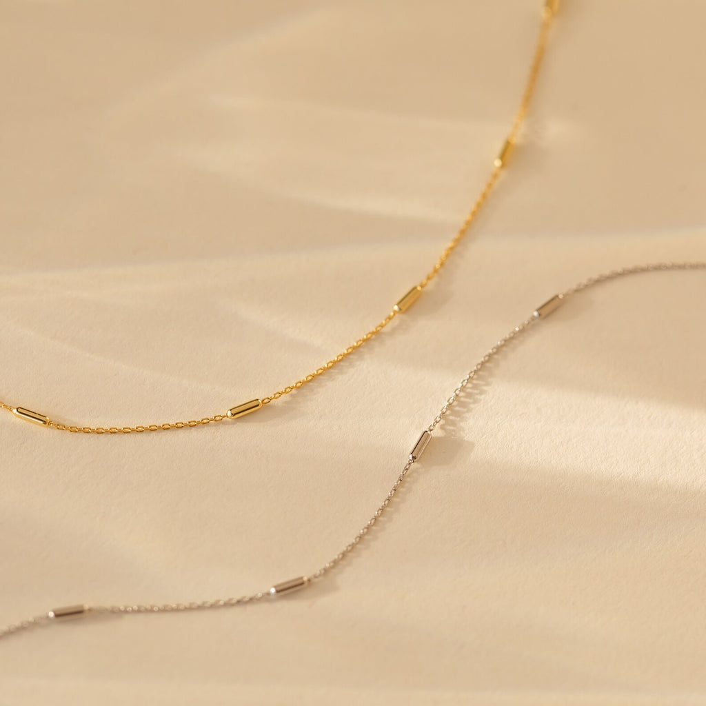 Bar Station Necklace with dainty charms and delicate chains, featuring small cylindrical beads, displayed on a beige background.