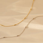 Bar Station Necklace with dainty charms and delicate chains, featuring small cylindrical beads, displayed on a beige background.