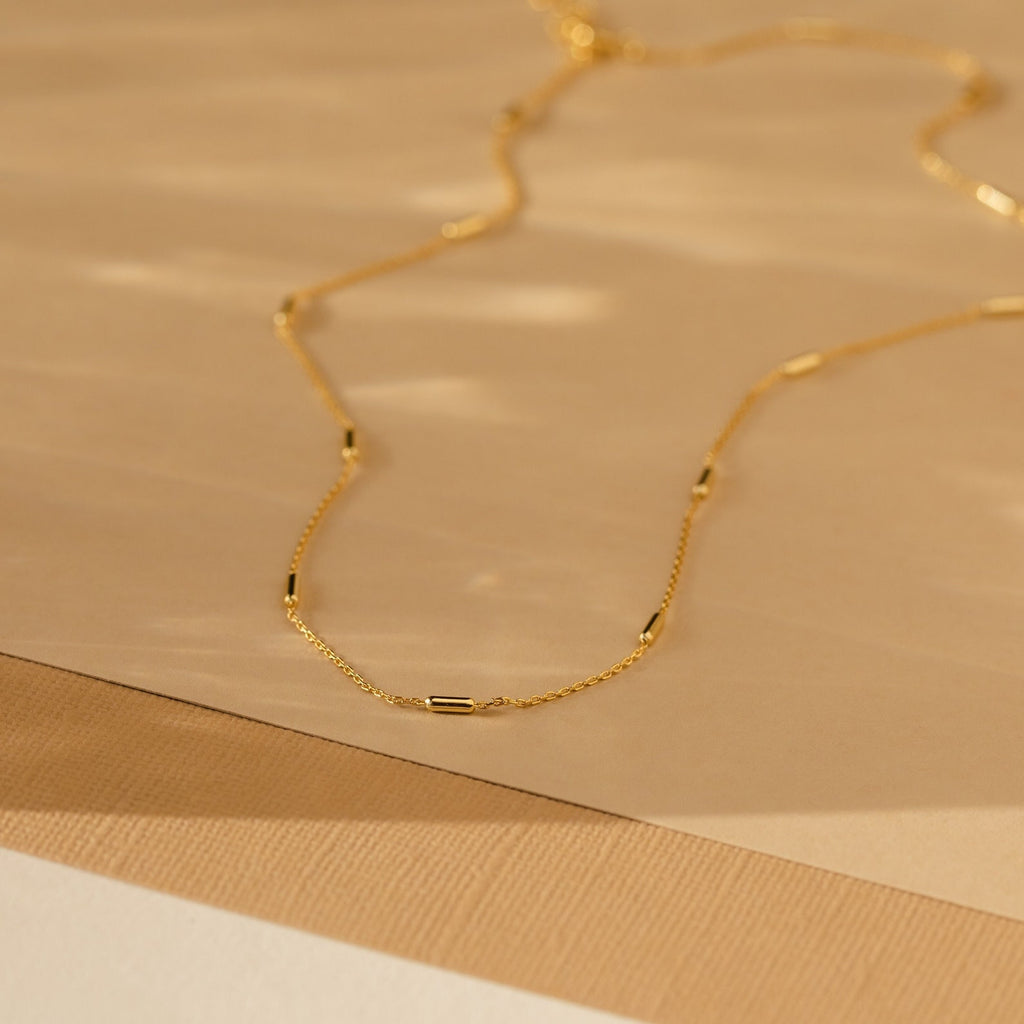Bar Station Necklace: A delicate gold necklace with small cylindrical beads on a fine chain, shown on a beige textured surface.
