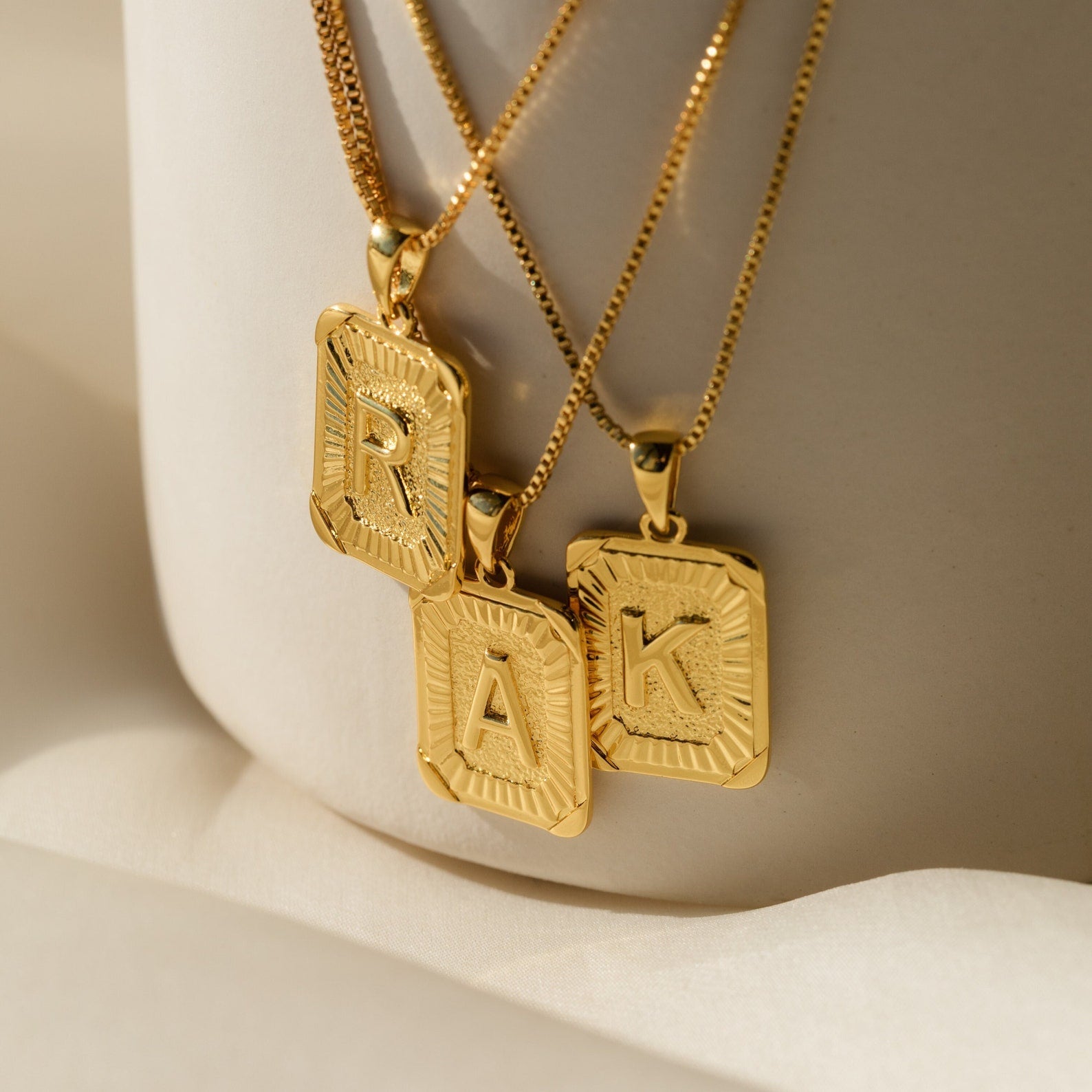 Gold initial pendant necklaces with rectangular engraved frames, featuring the letters R, A, and K on delicate box chains.
