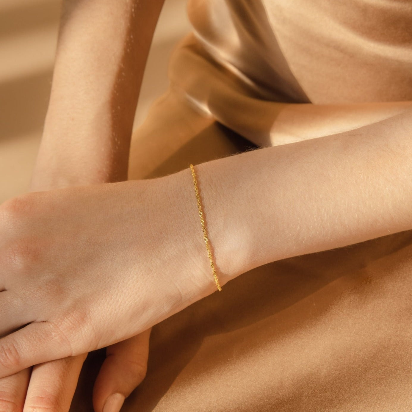 The Singapore Twist Bracelet adds a delicate touch to any wrist, beautifully complementing a beige satin dress—ideal for lovers of dainty, minimalist style.