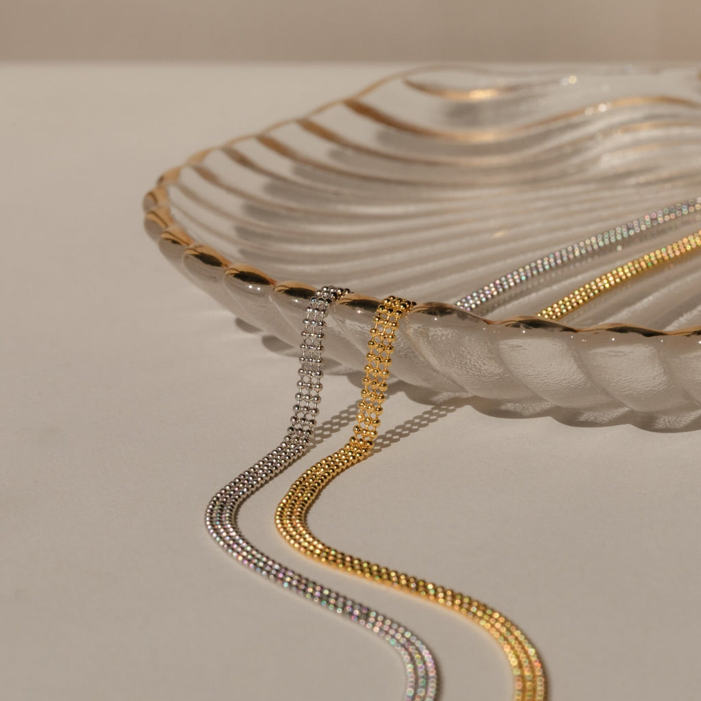 A clear, scalloped glass dish with gold edges displays the Tri-Stacked Beaded Necklace—an elegant, versatile piece—on a cream surface.