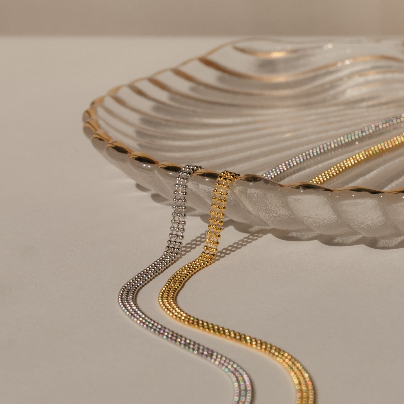 A clear, scalloped glass dish with gold edges displays the Tri-Stacked Beaded Necklace—an elegant, versatile piece—on a cream surface.