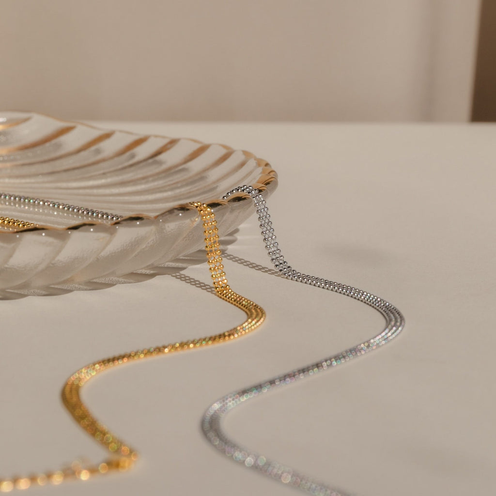 The Tri-Stacked Beaded Necklace, featuring gold and silver tones in a chunky minimalist style, is elegantly draped over the edge of a clear, shell-shaped glass dish.