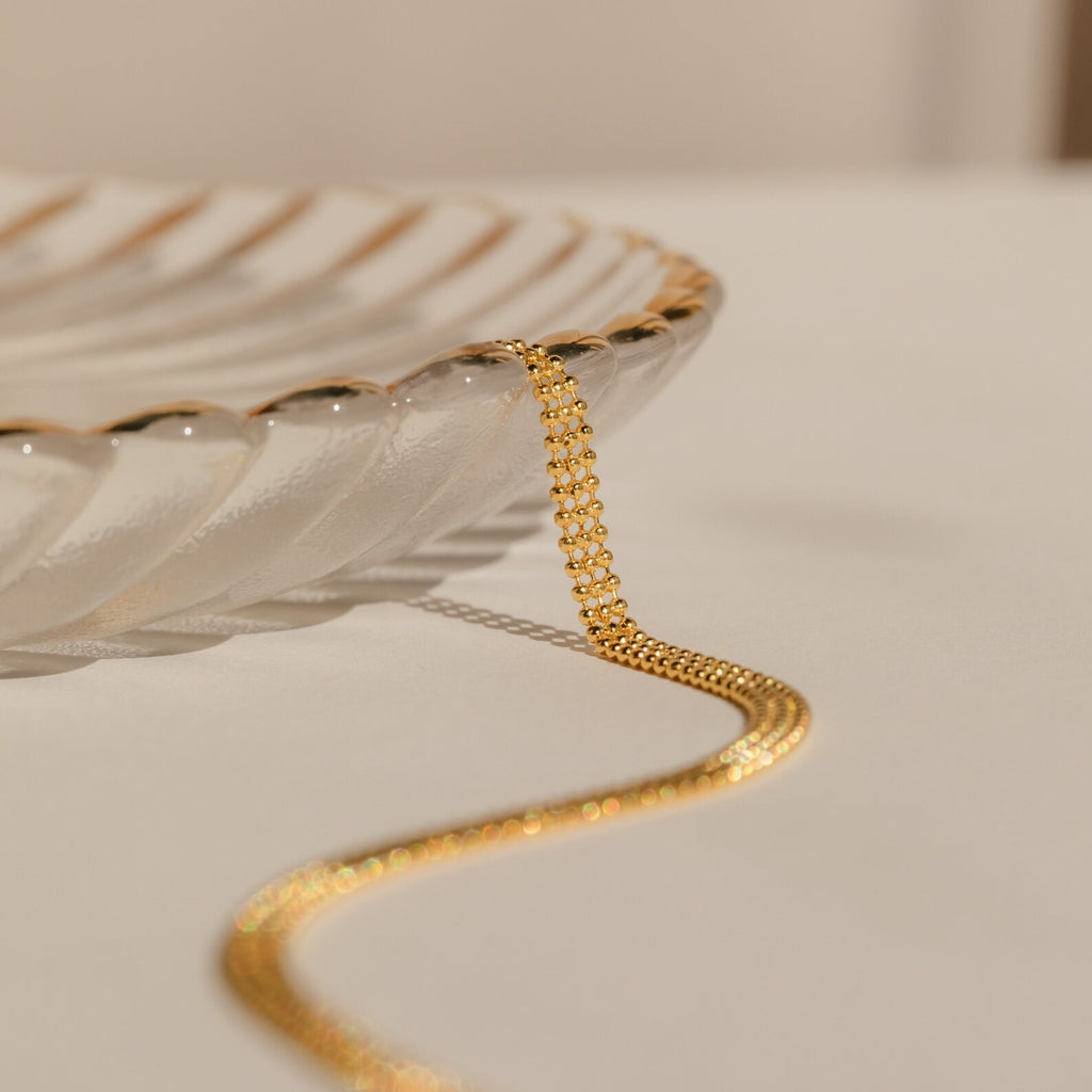 The Tri-Stacked Beaded Necklace, ideal for versatile jewelry styling, drapes over the edge of a clear, scalloped glass dish on a neutral surface.