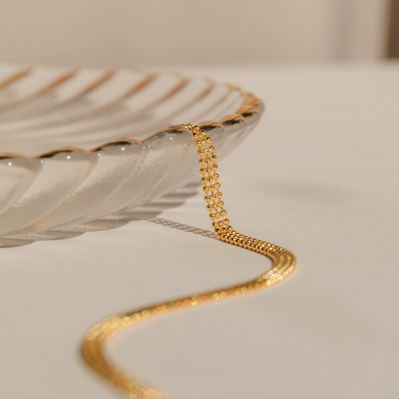 The Tri-Stacked Beaded Necklace, ideal for versatile jewelry styling, drapes over the edge of a clear, scalloped glass dish on a neutral surface.