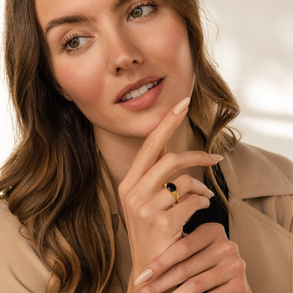 A woman with wavy brown hair in a beige coat wears the Dusk Signet Ring, touching her face thoughtfully to showcase this bold statement jewelry piece.