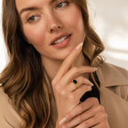 A woman with wavy brown hair in a beige coat wears the Dusk Signet Ring, touching her face thoughtfully to showcase this bold statement jewelry piece.