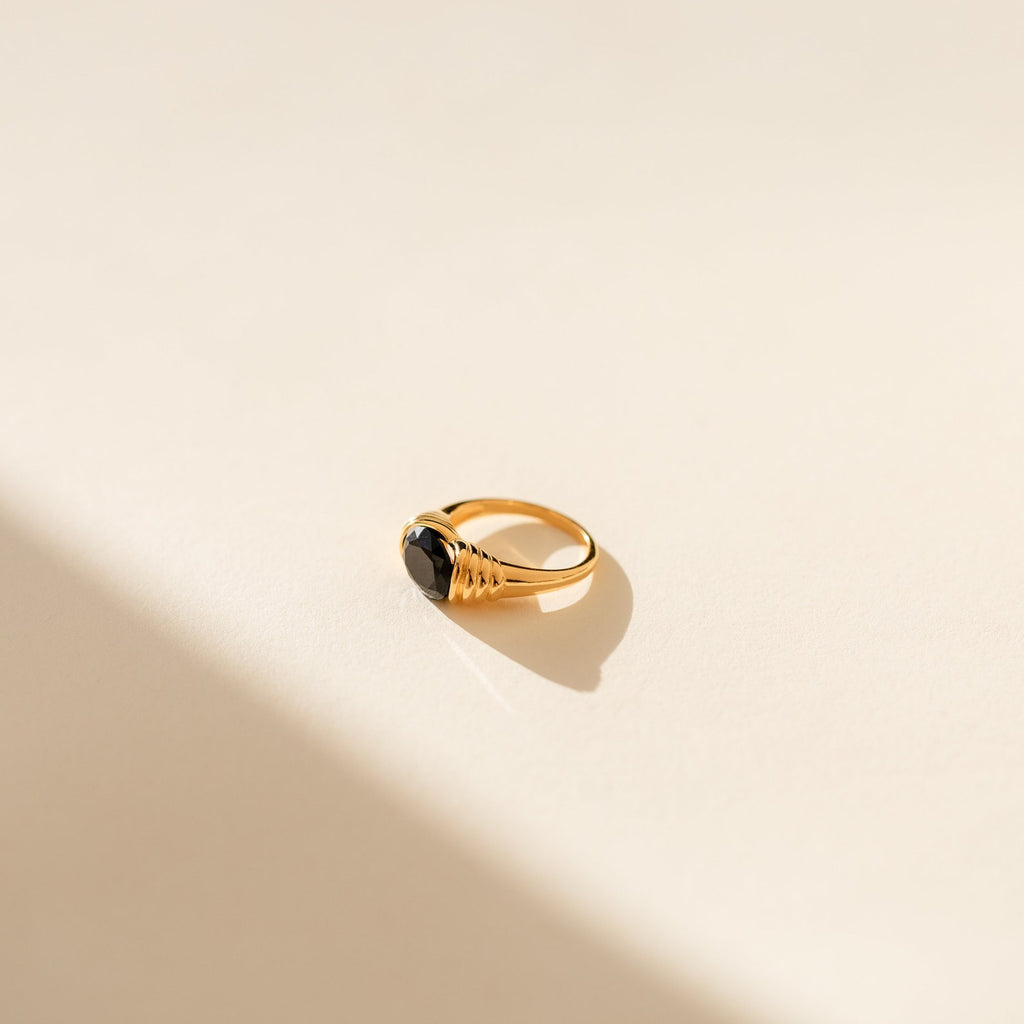 The Dusk Signet Ring is a gold vintage-inspired piece featuring a dark gemstone, showcased on a light neutral background with a soft shadow—an ideal statement jewelry accessory.