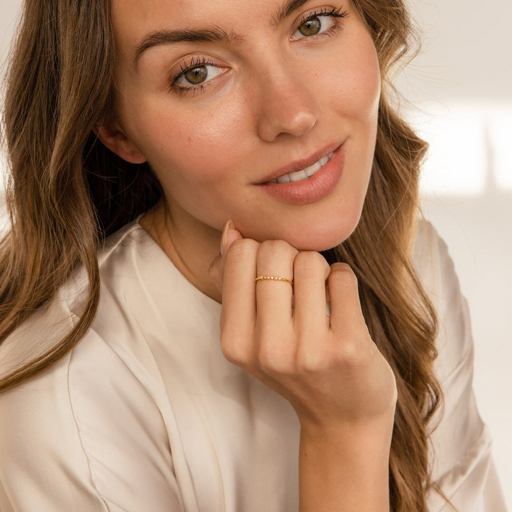 A woman with long brown hair smiles in a cream top, showcasing her Delicate Pearl Diamond Ring on her finger.