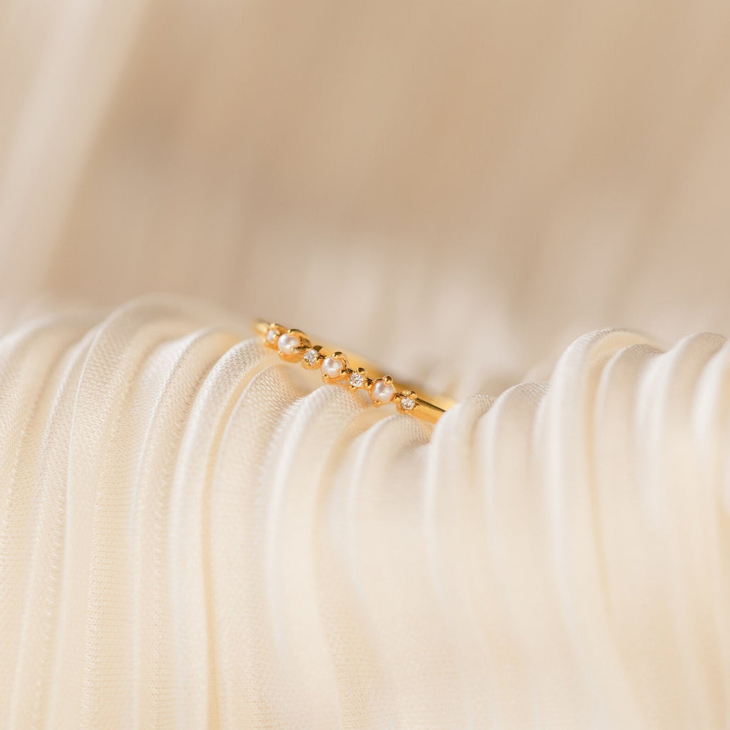 The Delicate Pearl Diamond Ring, featuring small pearls and diamonds in a gold band, rests on soft, pleated cream-colored fabric.