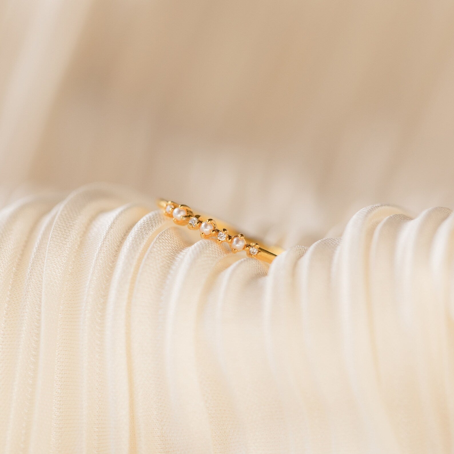 The Delicate Pearl Diamond Ring, featuring small pearls and diamonds in a gold band, rests on soft, pleated cream-colored fabric.