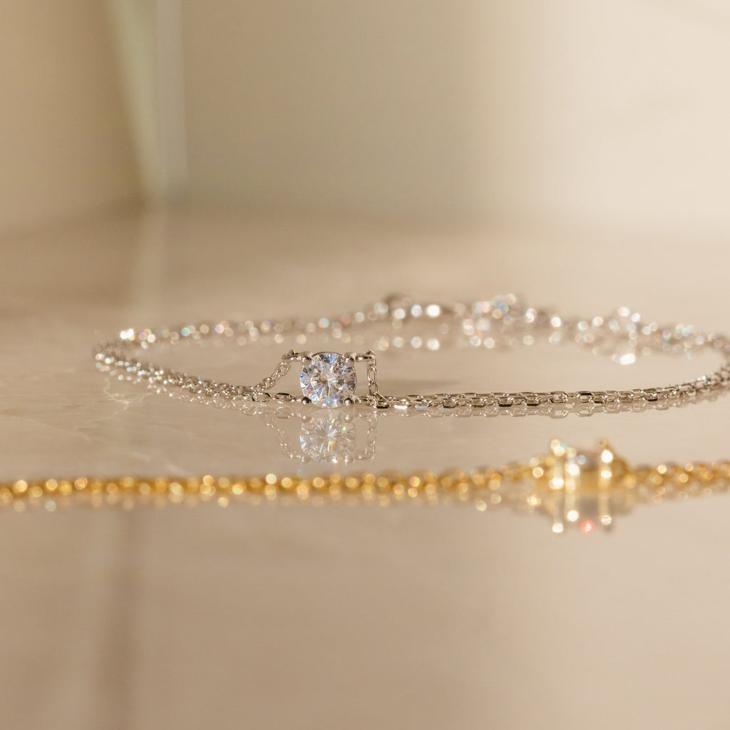The Duo Chain Diamond Bracelet features a sparkling gem and sleek gold double chain, elegantly displayed on a reflective surface—an ideal anniversary gift.