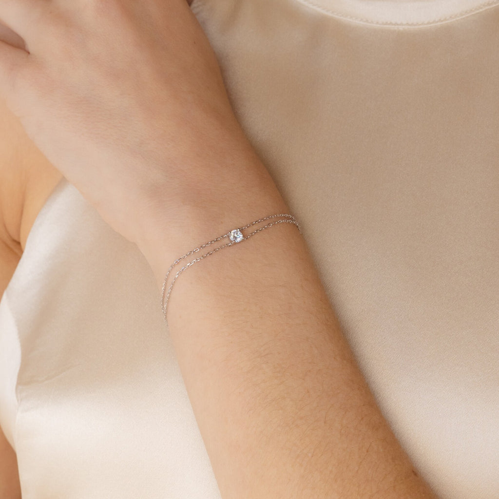 The Duo Chain Diamond Bracelet, featuring a subtle diamond on dual chains, elegantly accents the wrist over a cream satin top—a minimalist piece ideal as an anniversary gift.