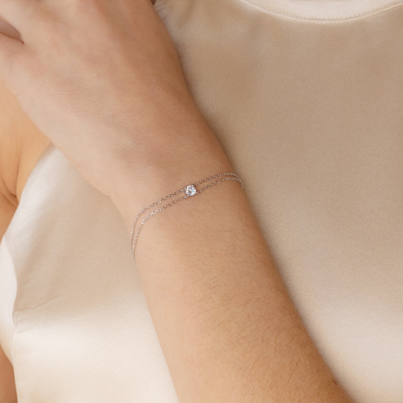 The Duo Chain Diamond Bracelet, featuring a subtle diamond on dual chains, elegantly accents the wrist over a cream satin top—a minimalist piece ideal as an anniversary gift.