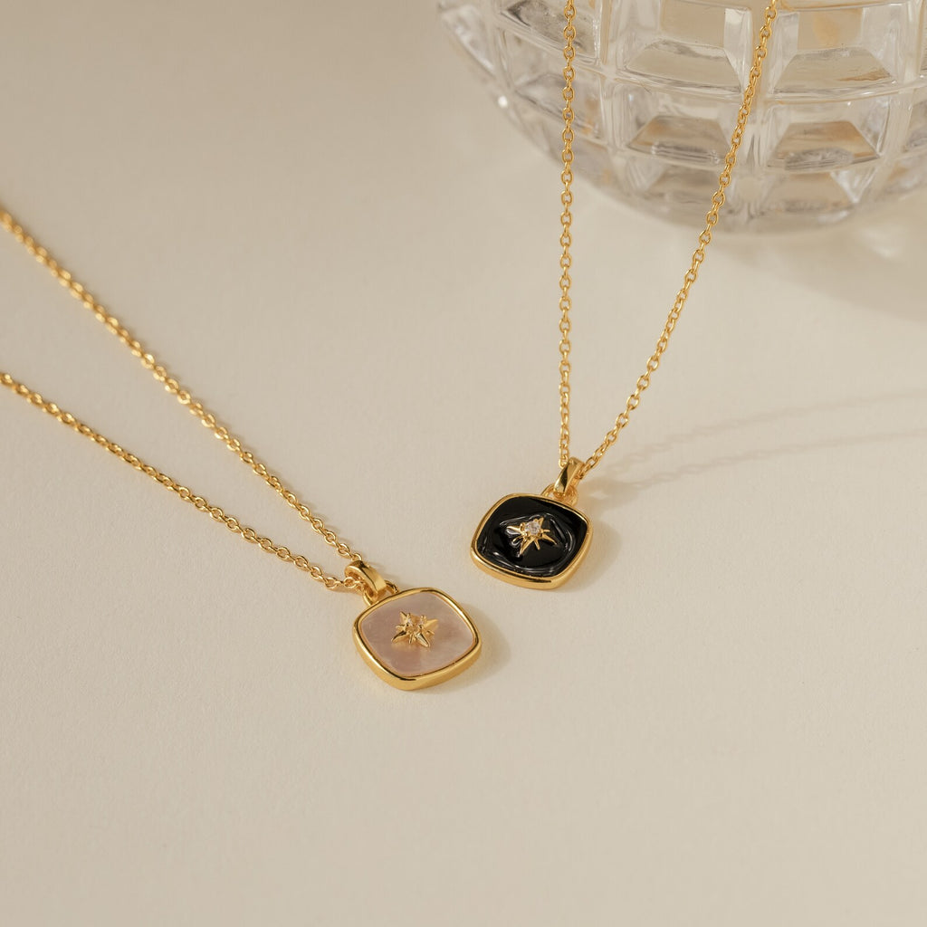 Two gold pendant necklaces—one with a pink stone and one with a black stone—rest on a cream surface, featuring the elegant Hermoine Pendant Necklace.