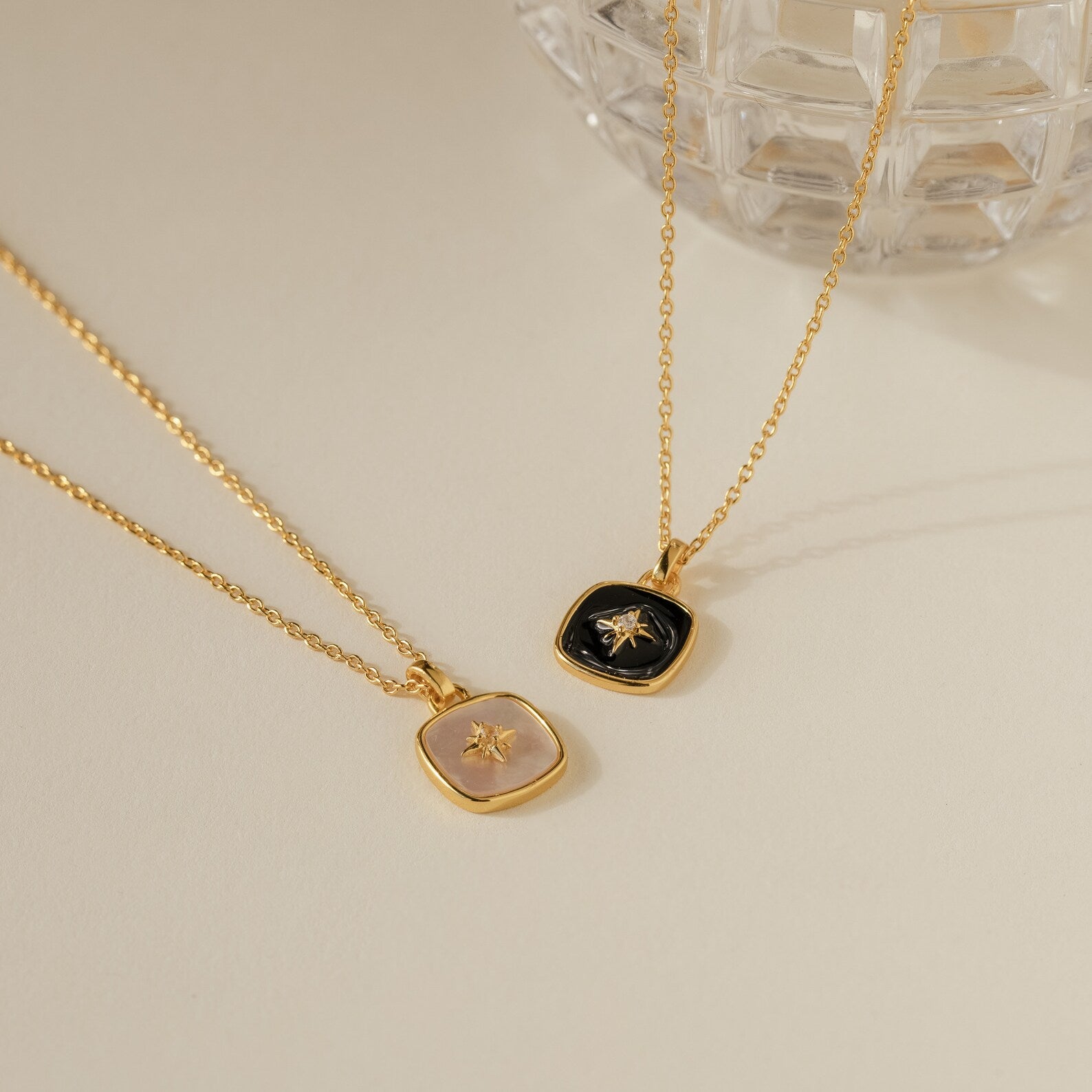 Two gold pendant necklaces—one with a pink stone and one with a black stone—rest on a cream surface, featuring the elegant Hermoine Pendant Necklace.