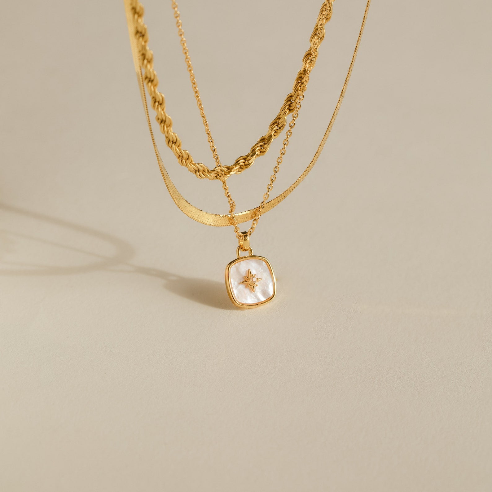 Three layered gold necklaces, featuring the Hermoine Pendant Necklace with a square star charm, elegantly arranged on a neutral background.