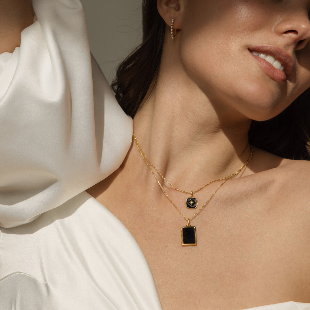 Woman in a white off-shoulder top layered with gold necklaces, featuring the eye-catching Hermoine Pendant Necklace with black pendants, paired with a gold hoop earring.
