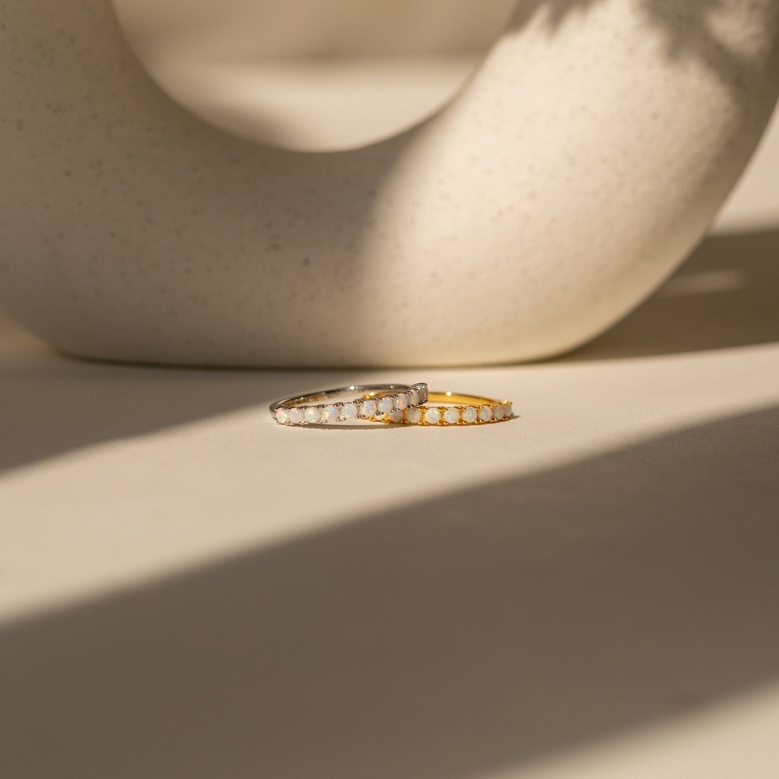 The Crescent Opal Ring, available in both silver and gold and accented with delicate opals, rests on a sunlit surface. Ideal as an anniversary gift or a meaningful promise ring to celebrate cherished moments.