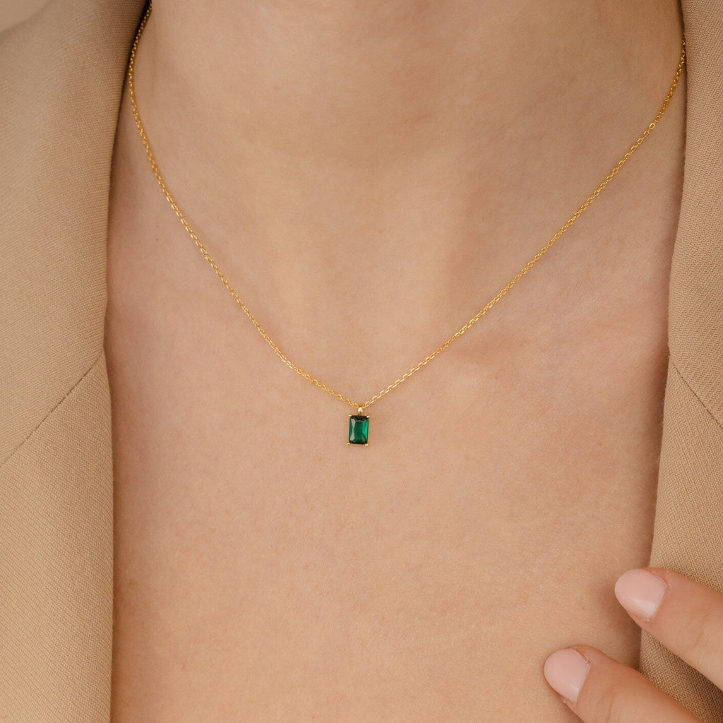 The Maia Emerald Necklace, featuring a small emerald pendant, rests elegantly on a person wearing a beige blazer.