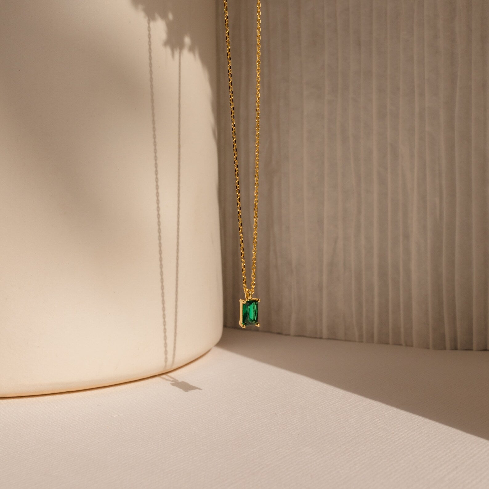 The Maia Emerald Necklace features a gold chain with a vibrant green emerald pendant, elegantly displayed against a soft, neutral background with gentle shadows.