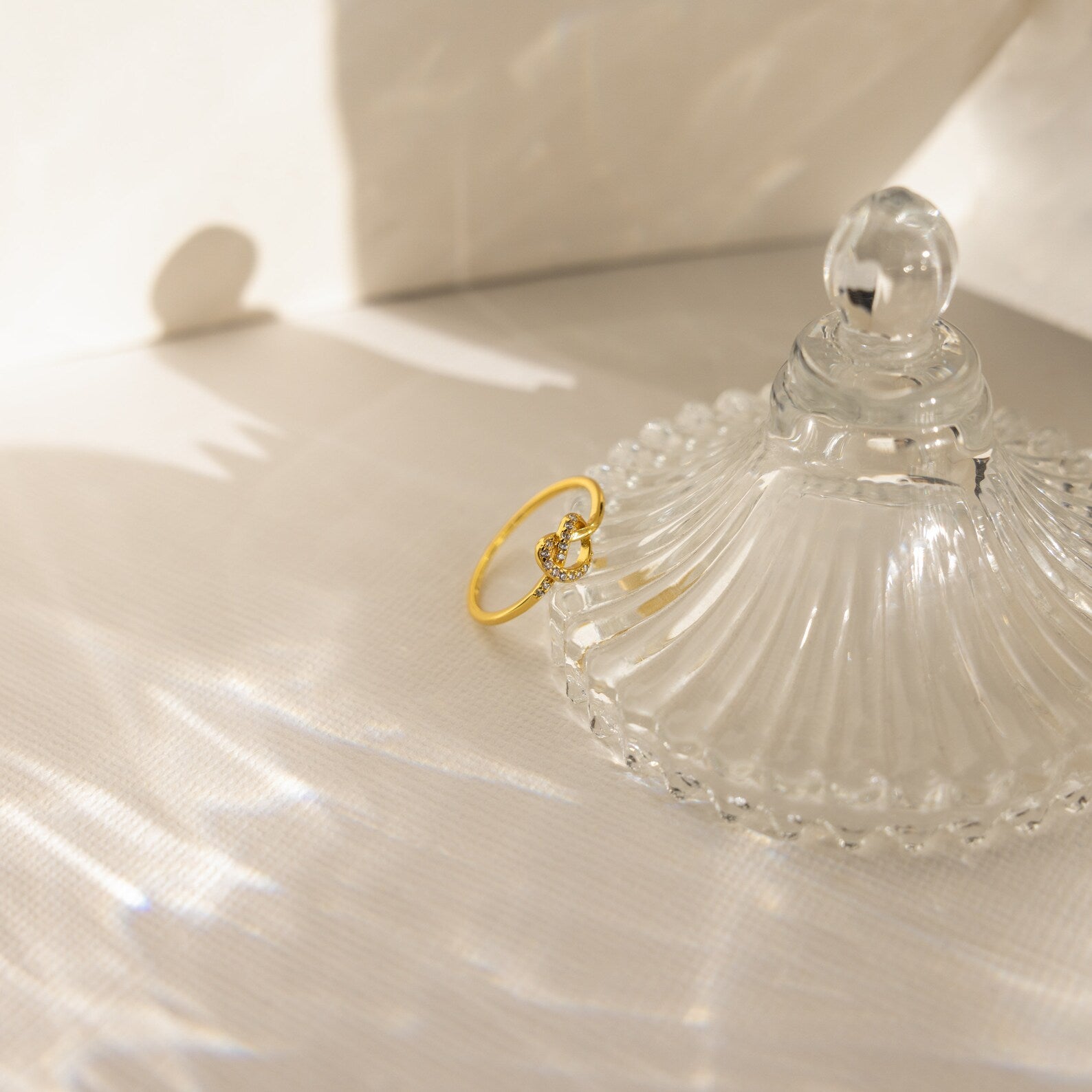 The Pave Heart Knot Ring gleams beside an ornate clear glass dish lid on a textured white surface—an elegant, romantic gift.