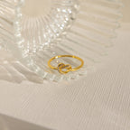 A Pave Heart Knot Ring rests against clear, textured glass on a light fabric surface—an elegant choice for a romantic gift or promise ring.