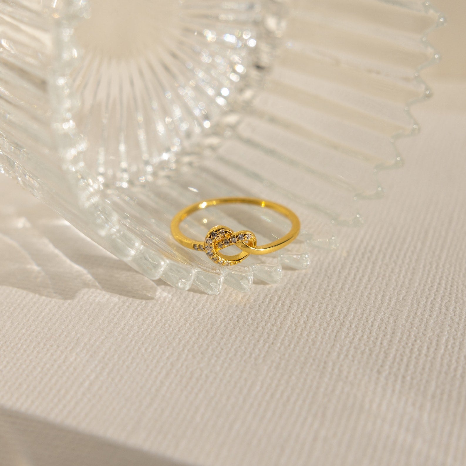 A Pave Heart Knot Ring rests against clear, textured glass on a light fabric surface—an elegant choice for a romantic gift or promise ring.