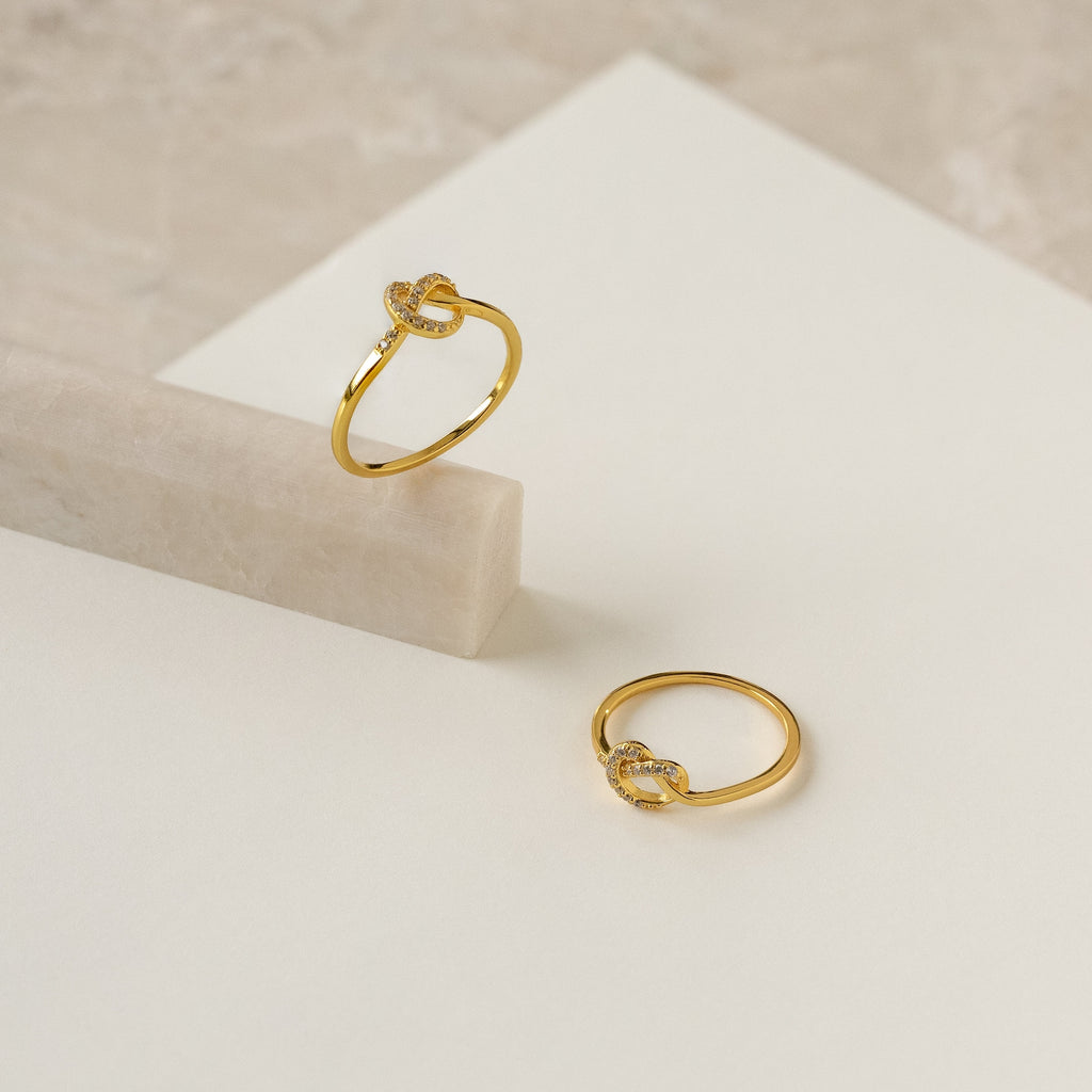 Two gold rings with small diamond accents, including a romantic Pave Heart Knot Ring, displayed on a white and marble background—perfect as a promise ring or meaningful romantic gift.
