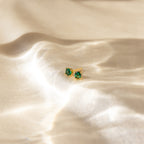 Booker Large Birthstone Studs—gold earrings featuring green gemstones—make a unique jewelry gift or personalized accessory, beautifully shown on soft, shimmery cream fabric with elegant folds.