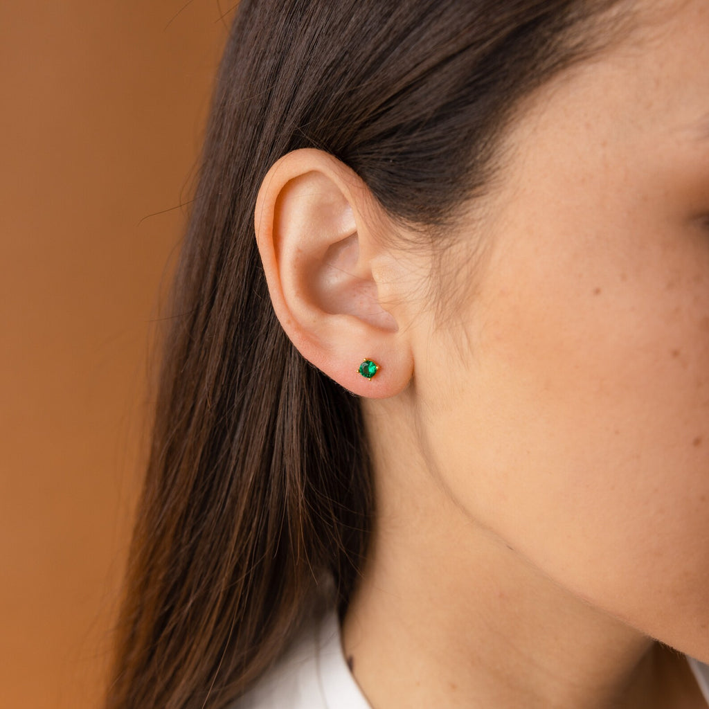 Close-up of a woman's ear wearing Booker Large Birthstone Studs in green against a light brown background, ideal as a thoughtful jewelry gift.