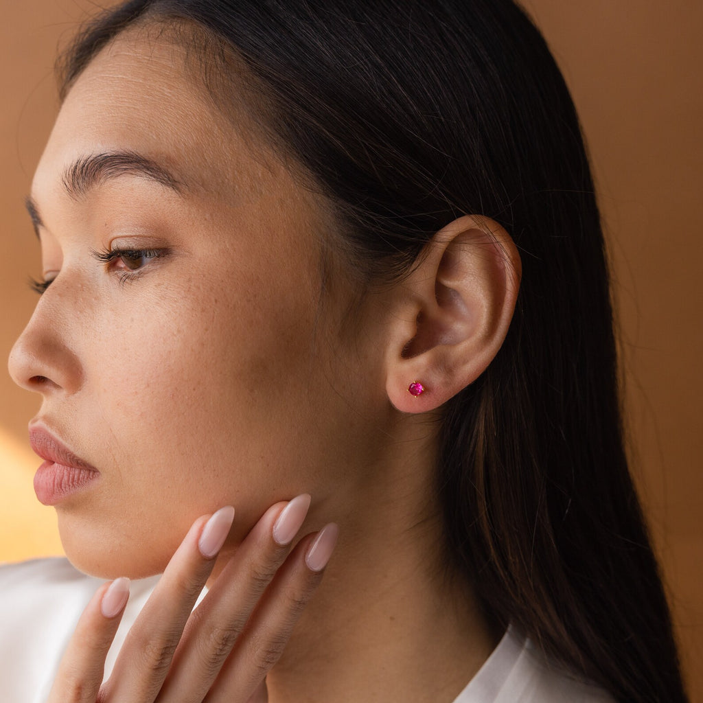 A woman with long dark hair wears Booker Large Birthstone Studs and touches her chin thoughtfully against a brown background—a perfect inspiration for a meaningful jewelry gift.