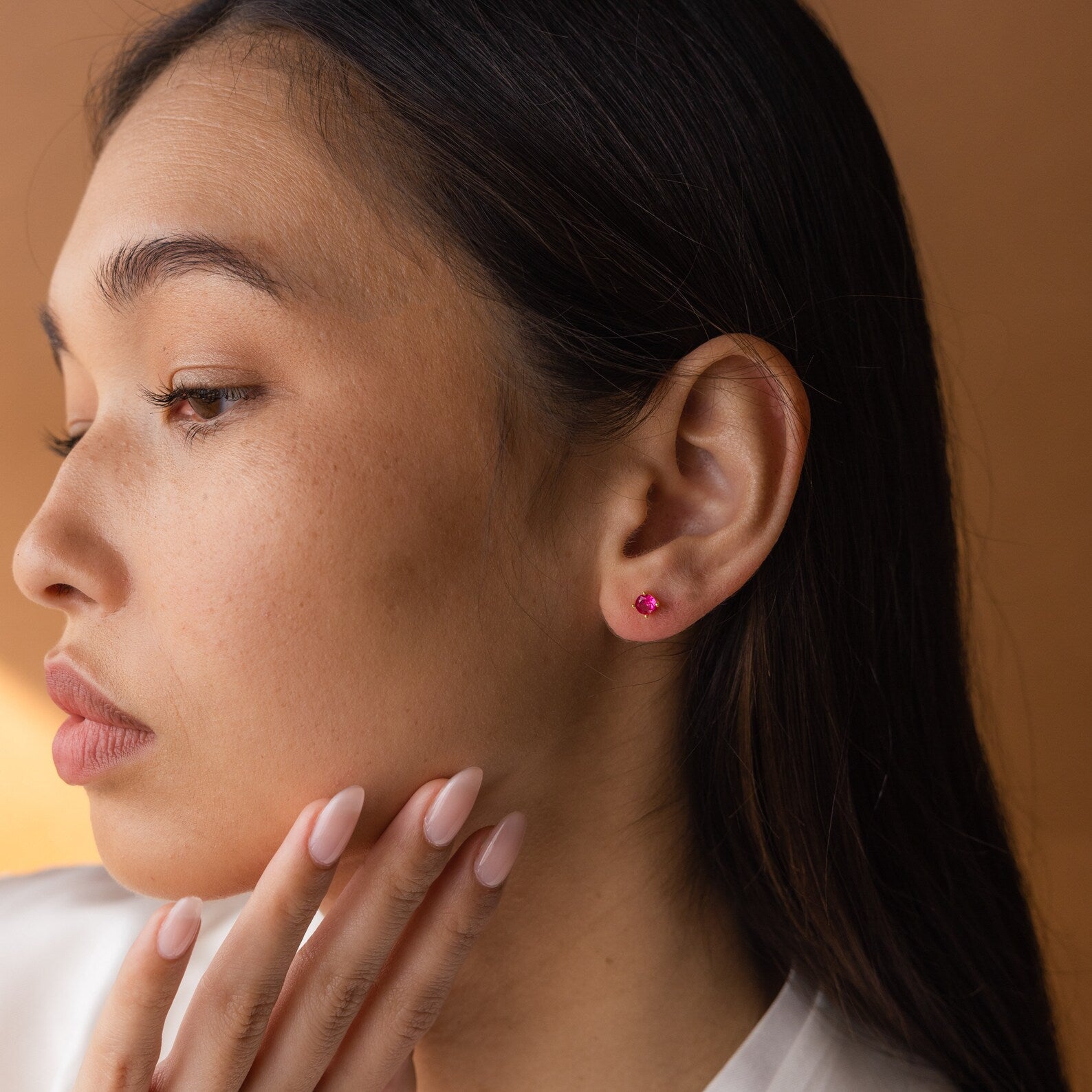 A woman with long dark hair wears Booker Large Birthstone Studs and touches her chin thoughtfully against a brown background—a perfect inspiration for a meaningful jewelry gift.