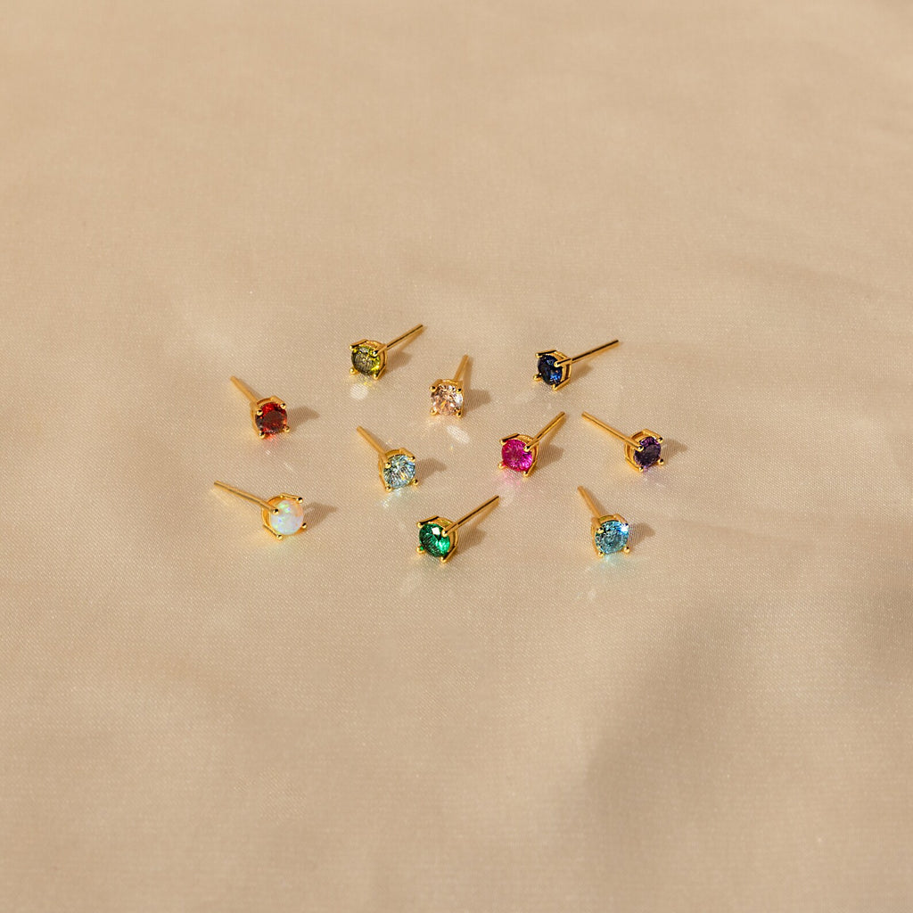 Nine Booker Large Birthstone Stud earrings, featuring colorful gemstones, are displayed on a beige fabric background.