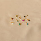 Nine Booker Large Birthstone Stud earrings, featuring colorful gemstones, are displayed on a beige fabric background.