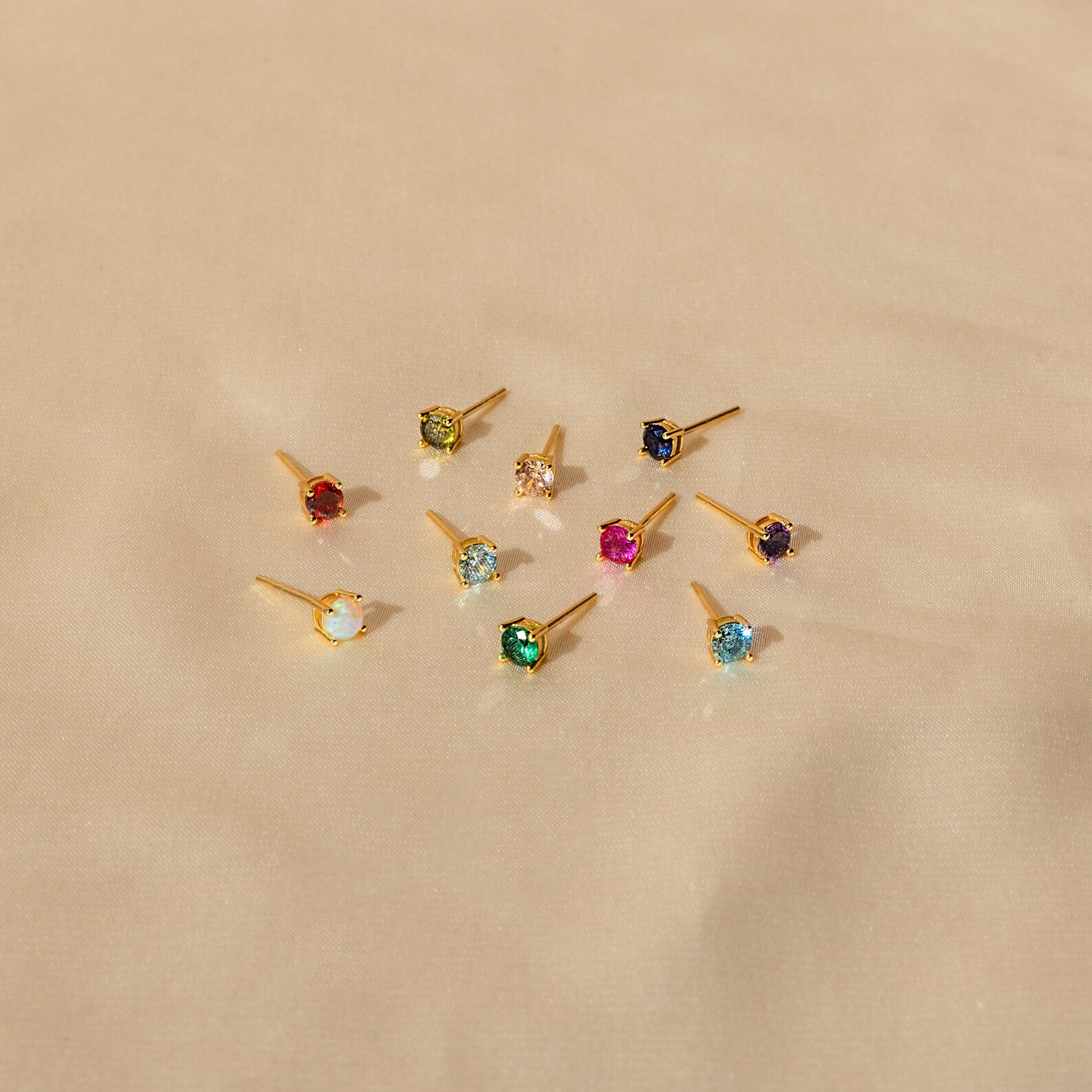 Nine Booker Large Birthstone Stud earrings, featuring colorful gemstones, are displayed on a beige fabric background.