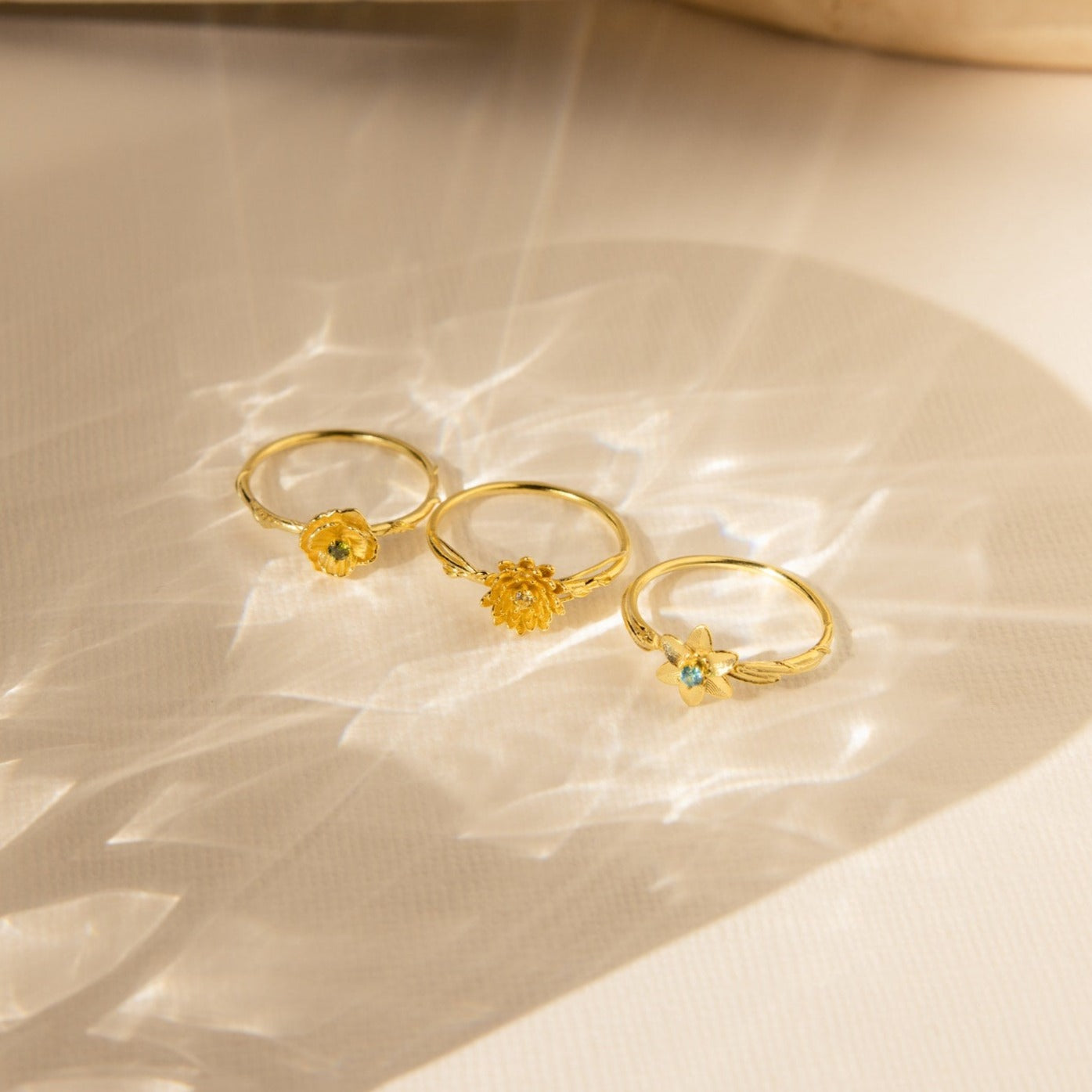Three Flower Birthstone Rings with yellow and blue gemstones rest on a light, textured surface with soft shadows.