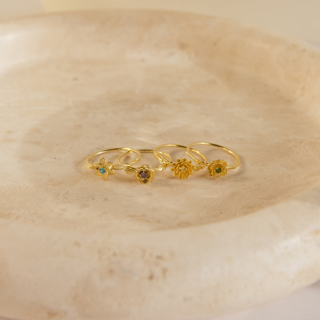 The Flower Birthstone Ring, a delicate gold ring featuring a small floral design with vintage inspiration, is displayed on a light beige, round stone dish.