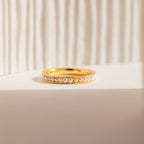 The Alicia Pearl Inlay Band features a row of small pearls set on gold, displayed on a textured white background. Ideal as a stackable ring, it offers minimalist style and understated elegance.