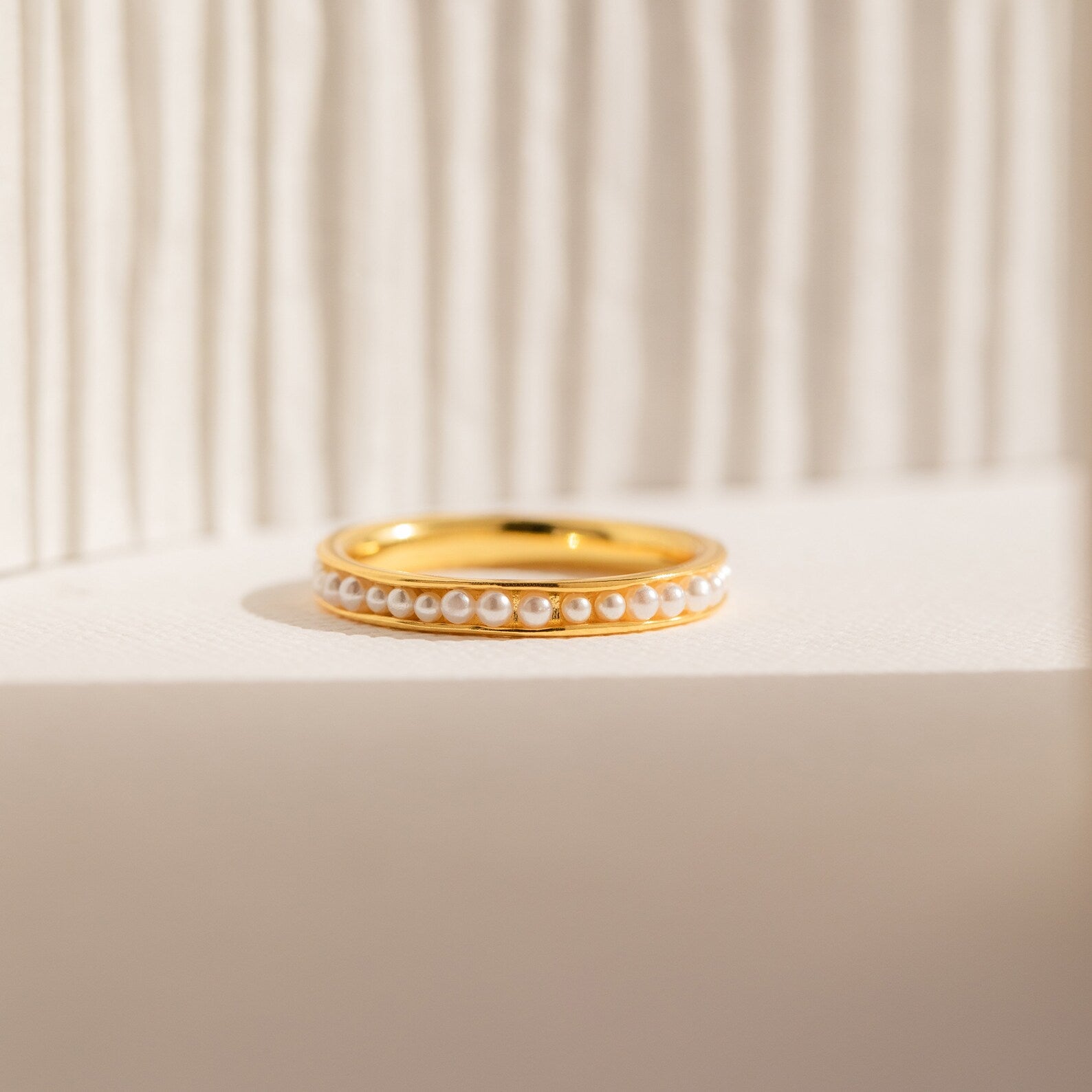 The Alicia Pearl Inlay Band features a row of small pearls set on gold, displayed on a textured white background. Ideal as a stackable ring, it offers minimalist style and understated elegance.