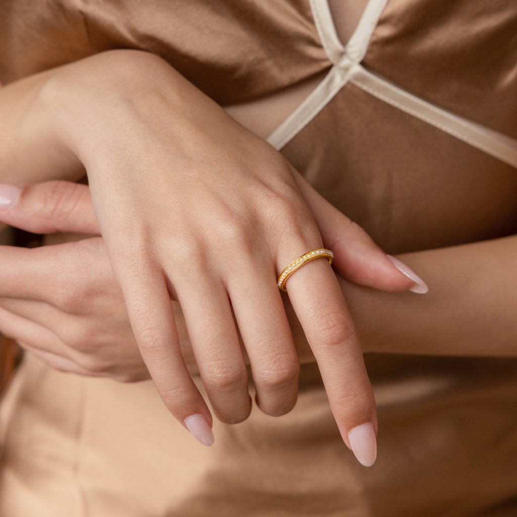 Close-up of a woman's hand wearing the Alicia Pearl Inlay Band, a gold stackable ring with pearl details, against a beige satin dress.